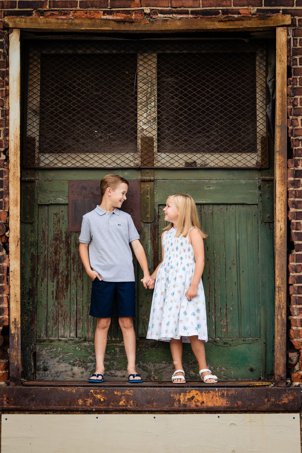Two children holding hands in front of a rustic green door, boy in gray polo and girl in floral dress smiling at each other.