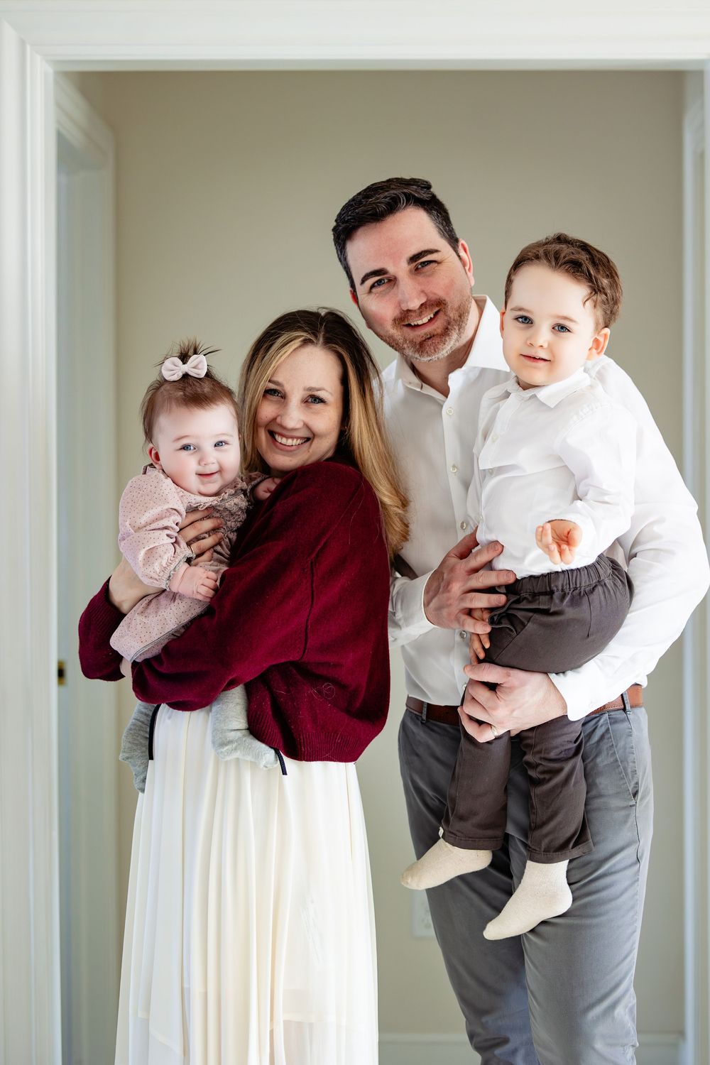 A smiling family of four poses indoors, parents holding a baby girl and toddler boy, dressed in elegant white and red attire.