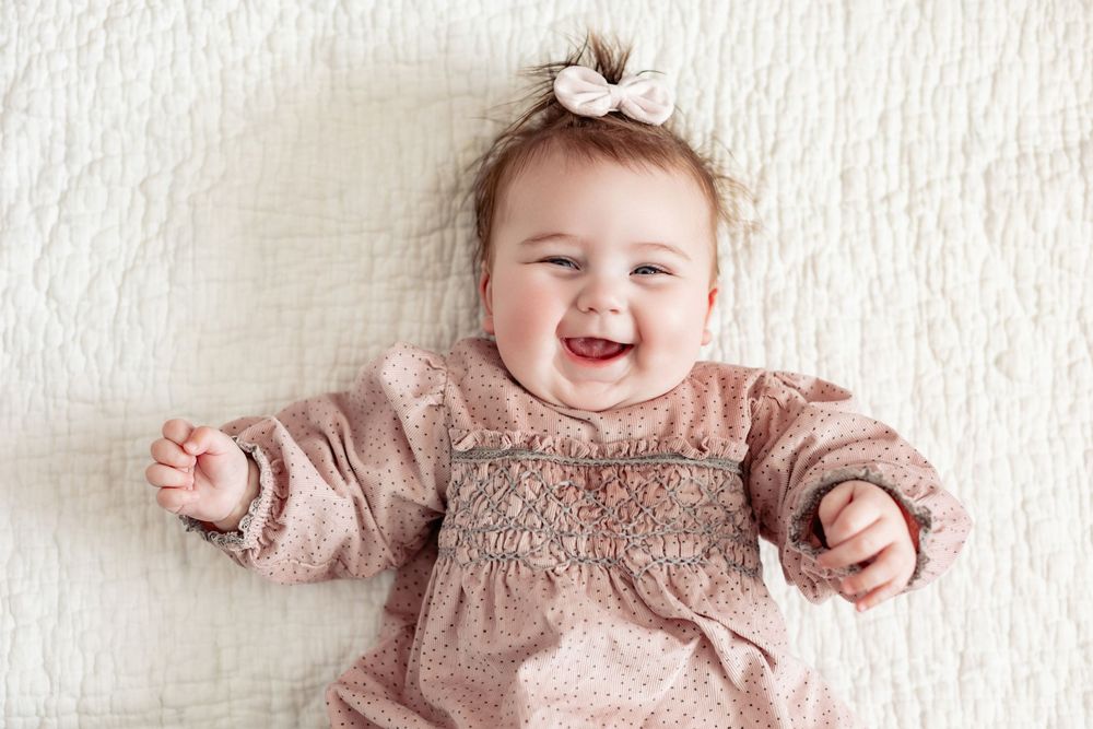 Happy baby girl in pink dress lying on white blanket, smiling joyfully with a bow in her hair.