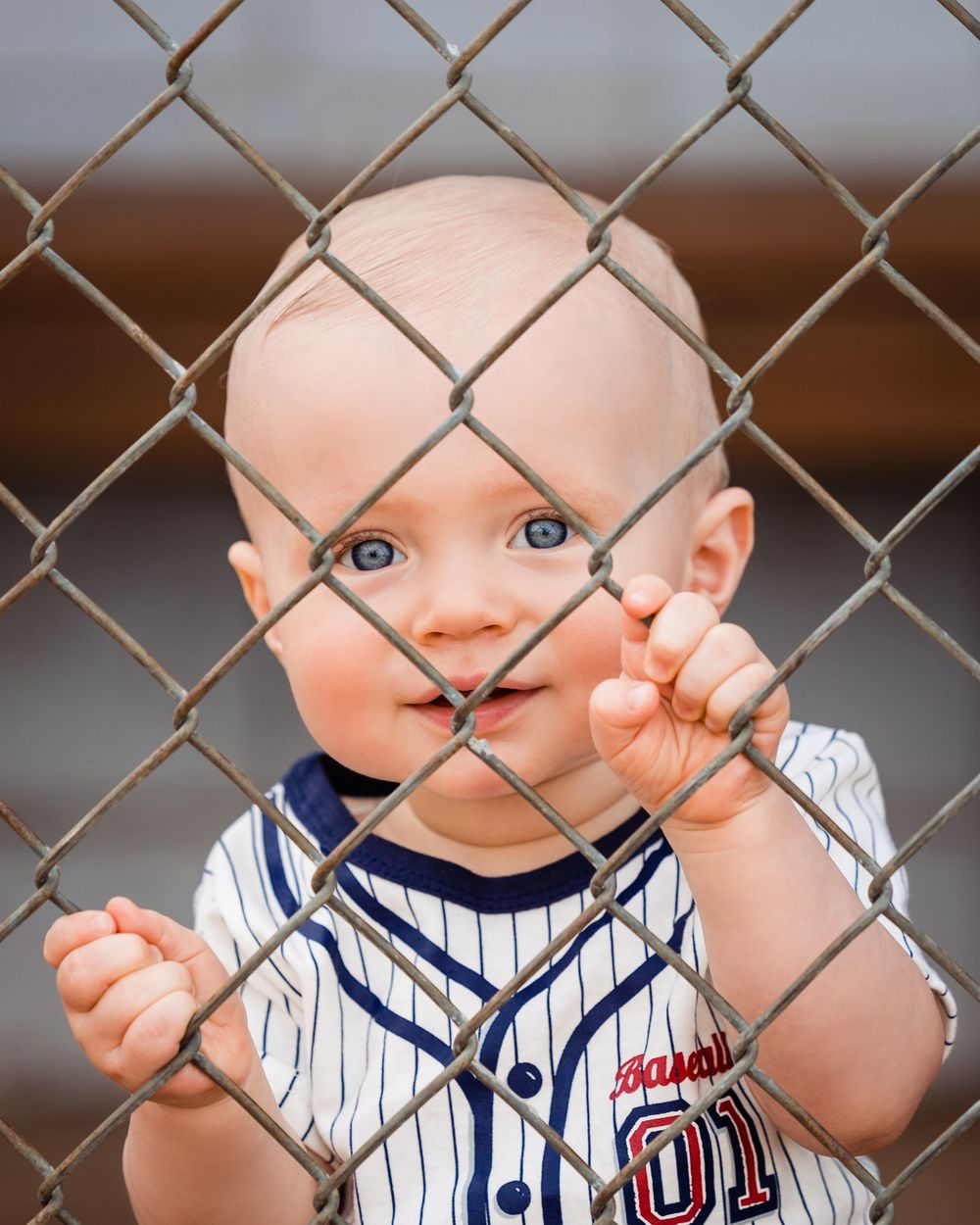 Adorable baby in a baseball jersey gripping a chain-link fence with bright blue eyes and a cheerful smile.