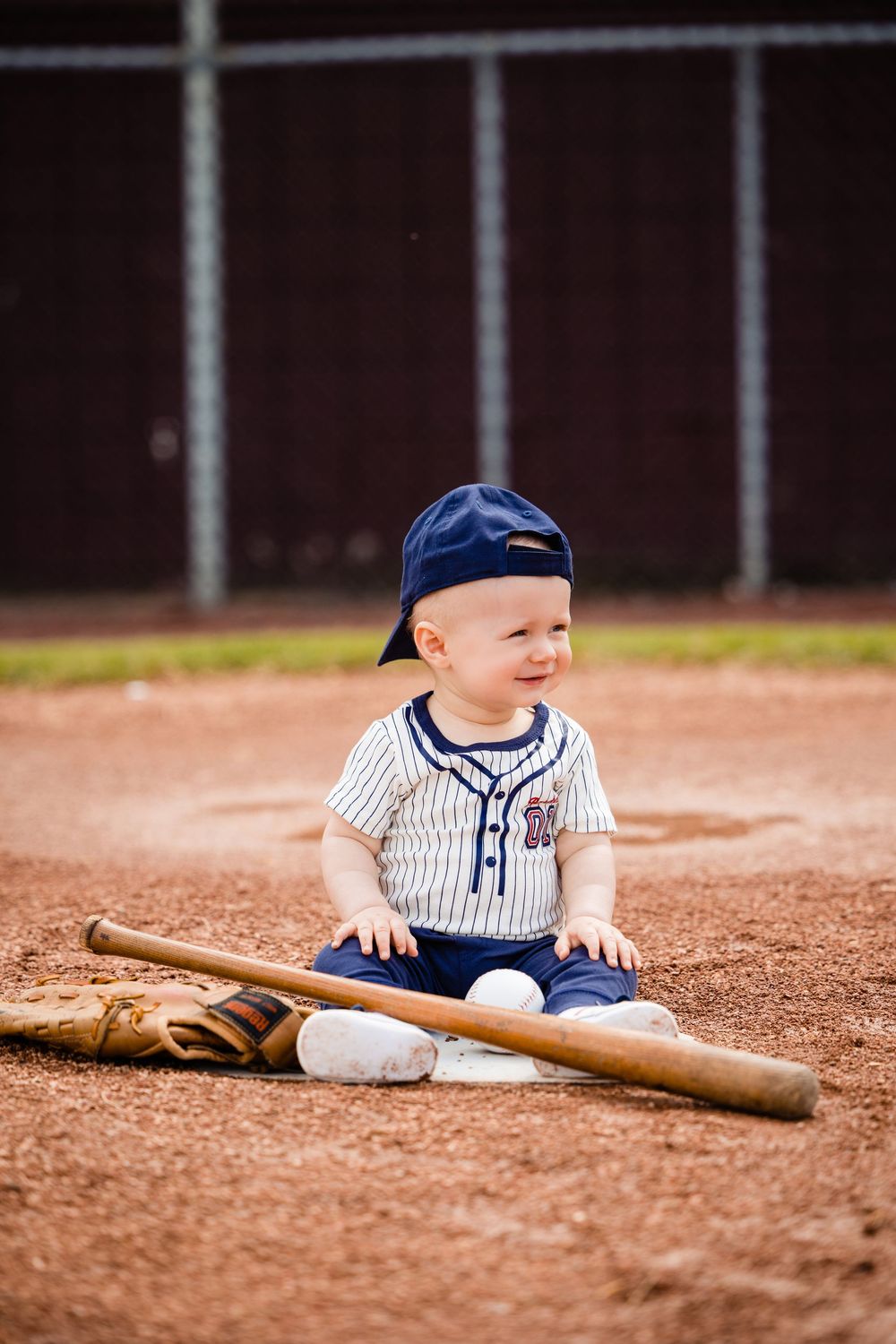 Adorable baby in baseball uniform and cap sits on dirt field with wooden bat and glove.