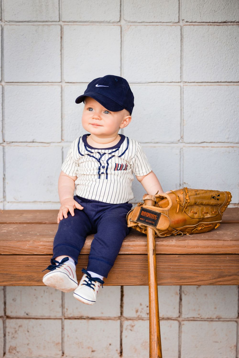 Adorable baby in baseball uniform with Nike cap, mitt, and bat sitting on dugout bench.