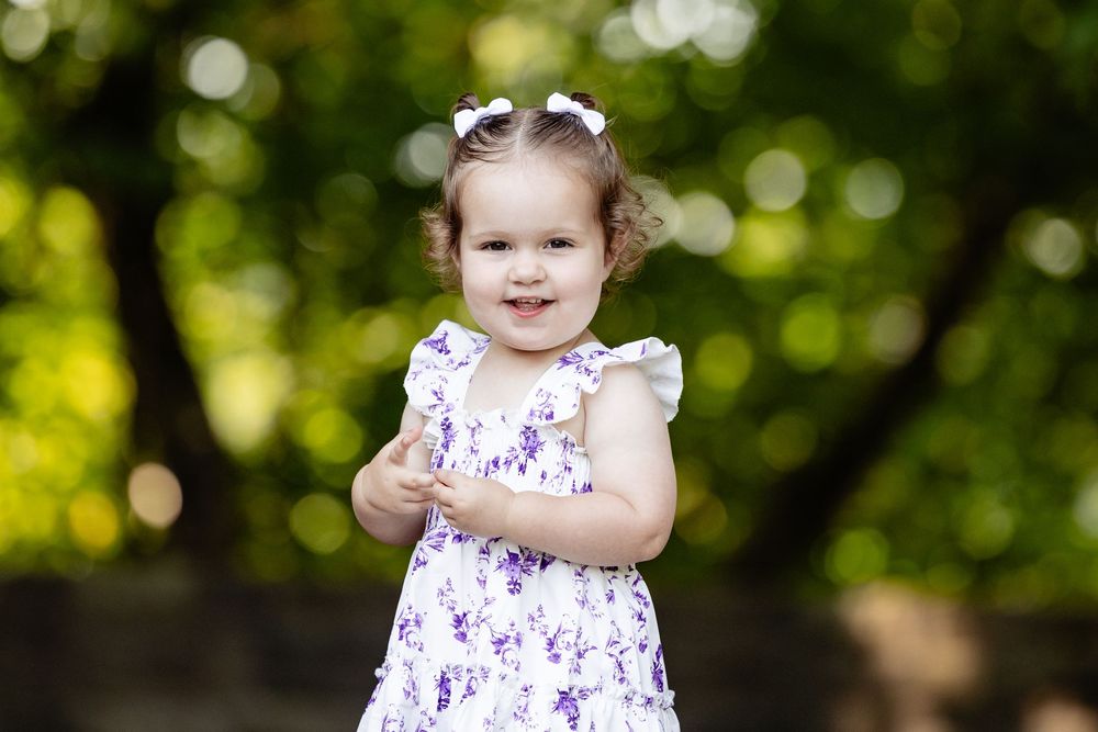 A smiling toddler girl in a white floral dress with purple flowers poses outdoors with green bokeh background.