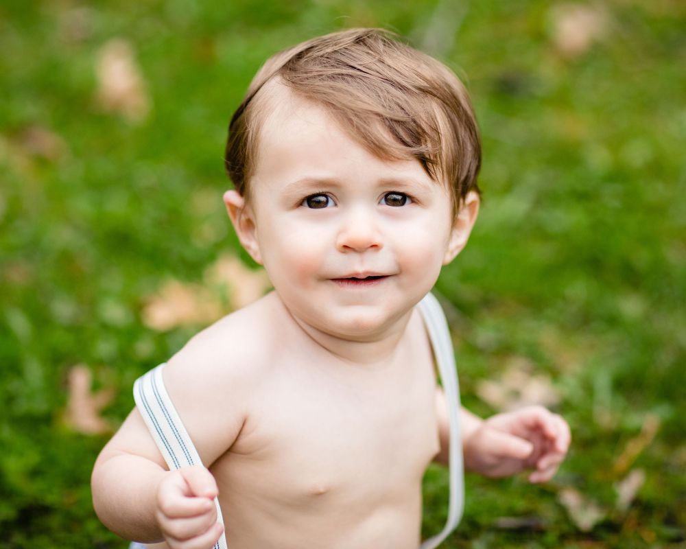 Cute toddler boy with brown hair smiling outdoors on green grass, shirtless with white straps.