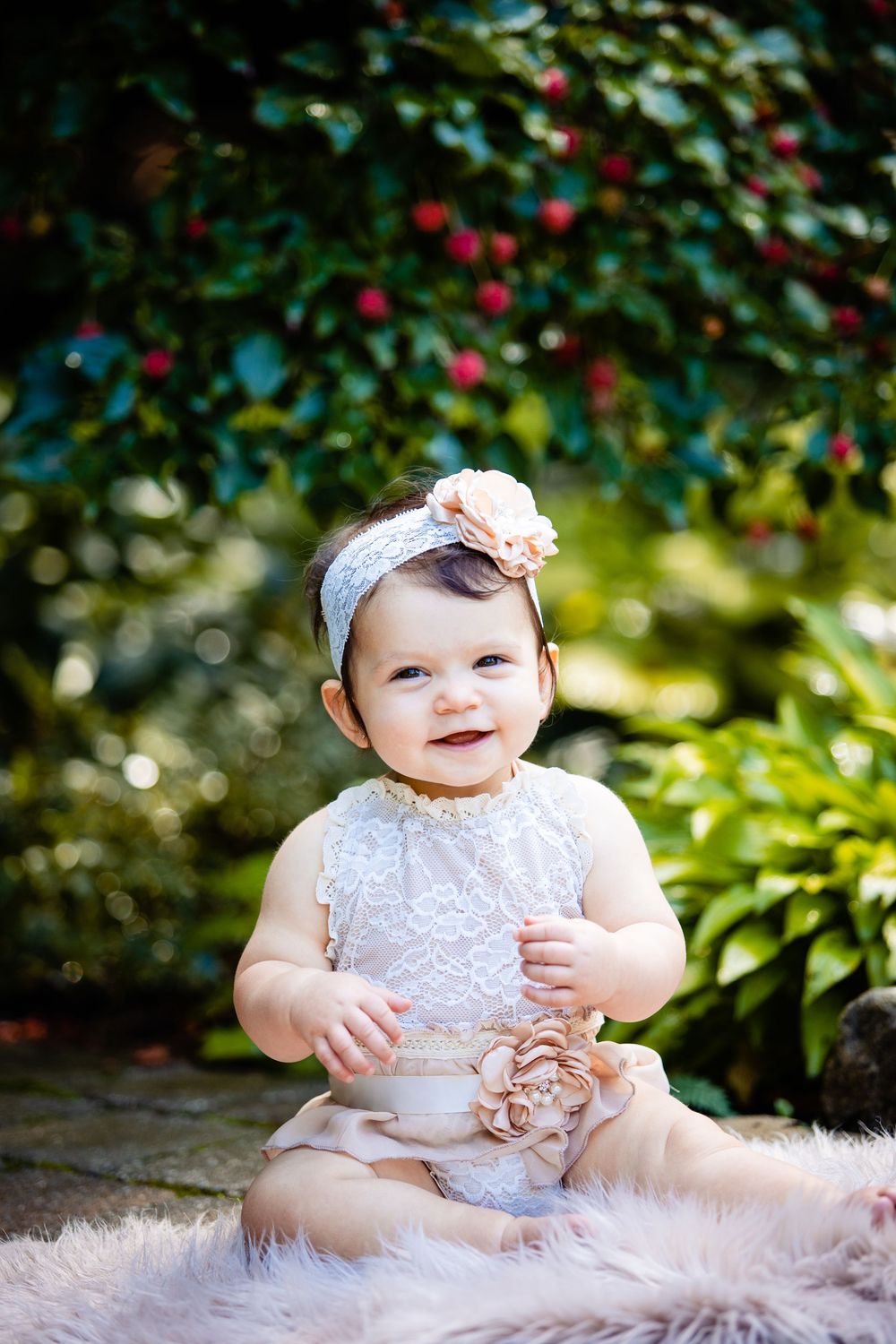 Smiling baby girl in white lace outfit and floral headband sitting outdoors on fur rug among lush green garden.