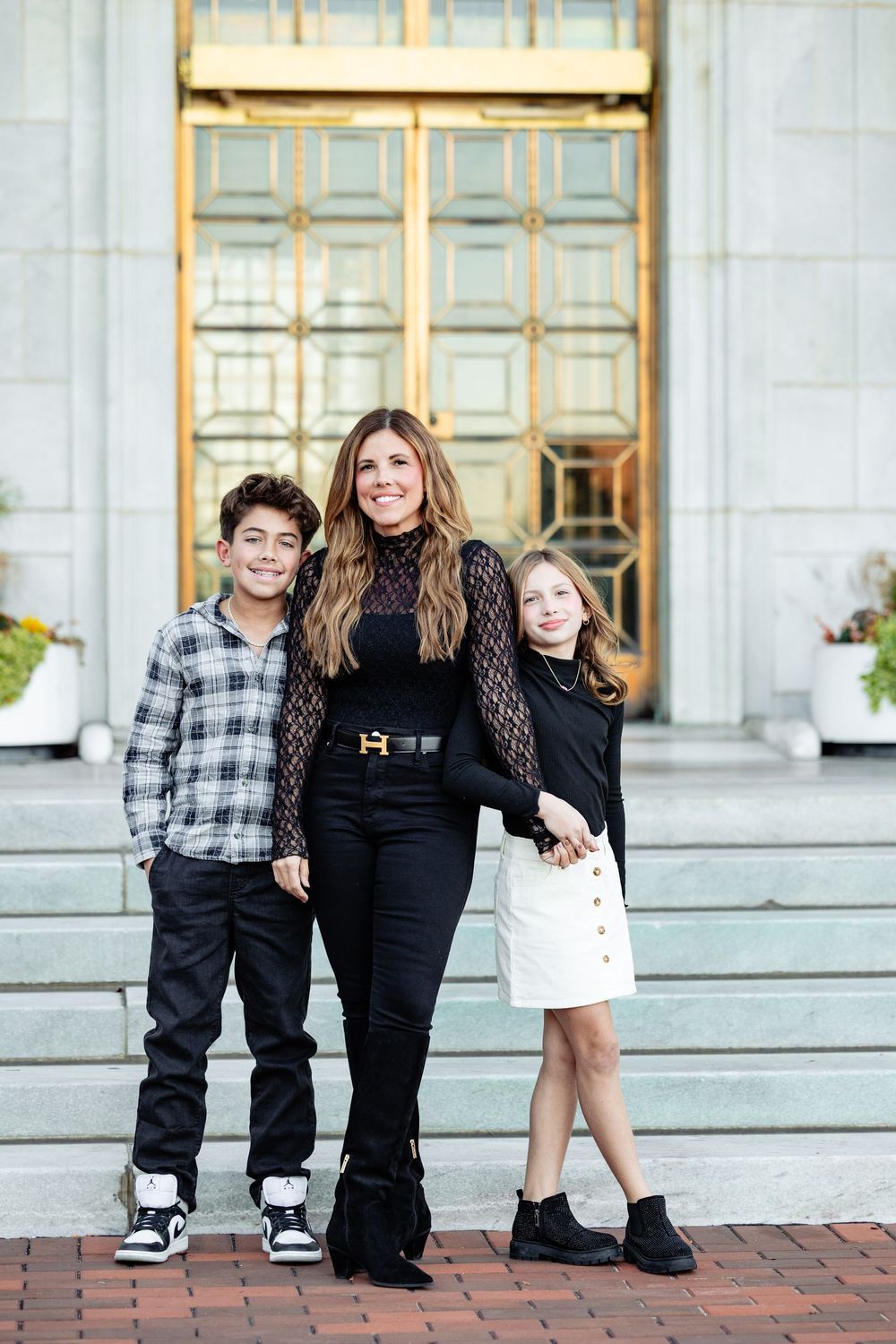 A woman in black outfit poses with two children on brick steps before golden doors.