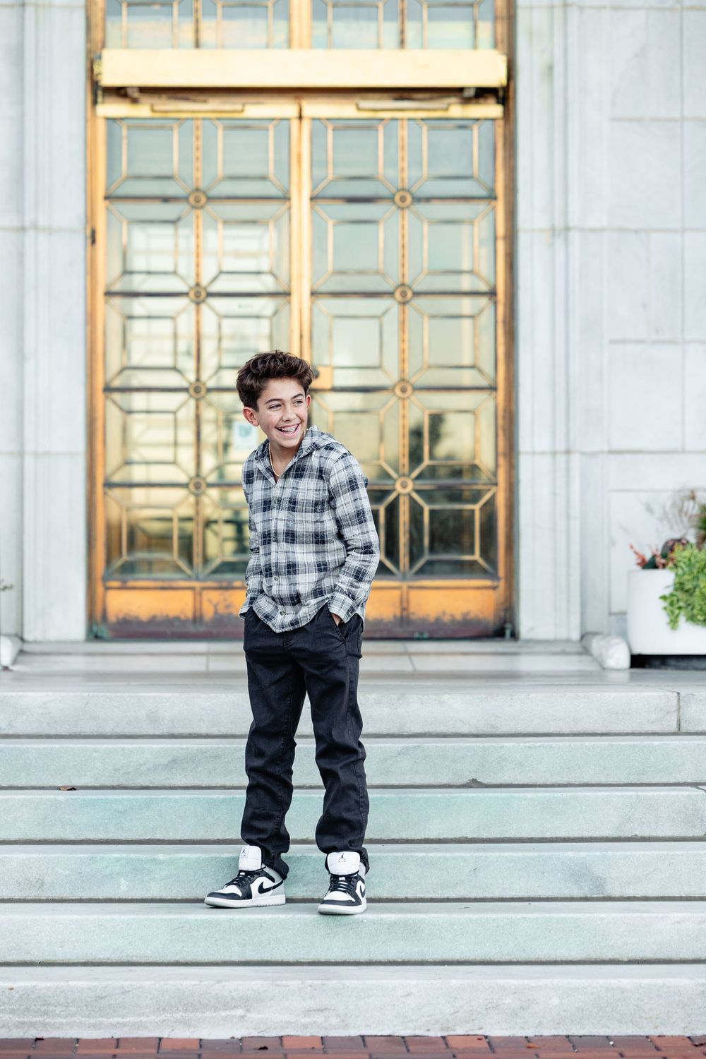 Smiling teenage boy in plaid shirt and black jeans standing on steps in front of ornate golden doors.
