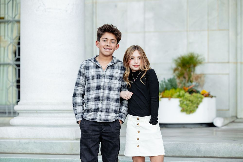 Two stylish kids posing outdoors, boy in plaid shirt and girl in white skirt with black top near white columns.