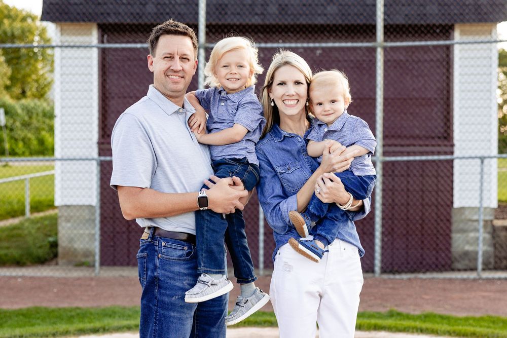 Happy family of four in matching blue outfits smiling outdoors near a baseball field on a sunny day.