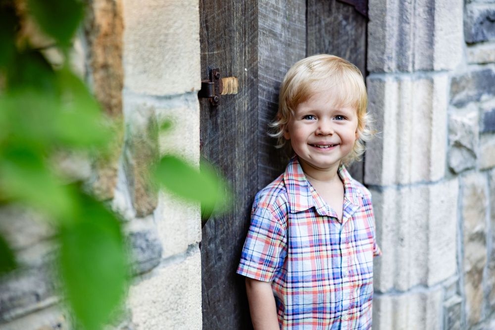 Smiling toddler boy in plaid shirt standing by rustic wooden door and stone wall outdoors.