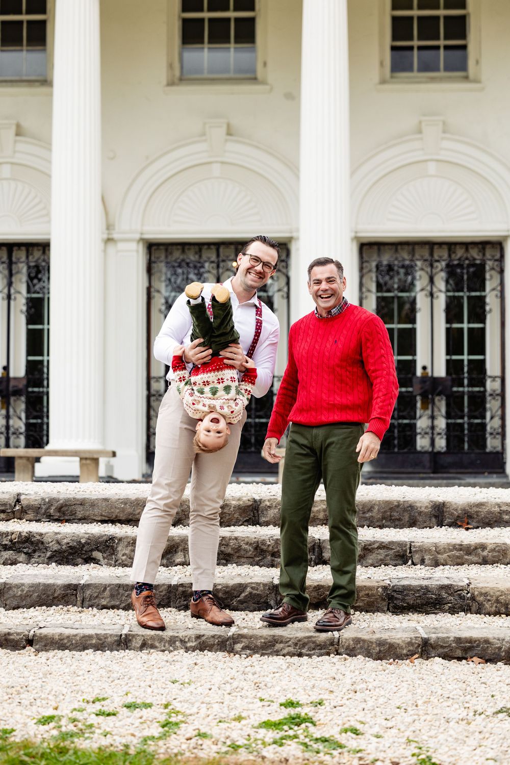 Two men smiling on steps of white building, one holding a child upside down, wearing festive holiday sweaters.