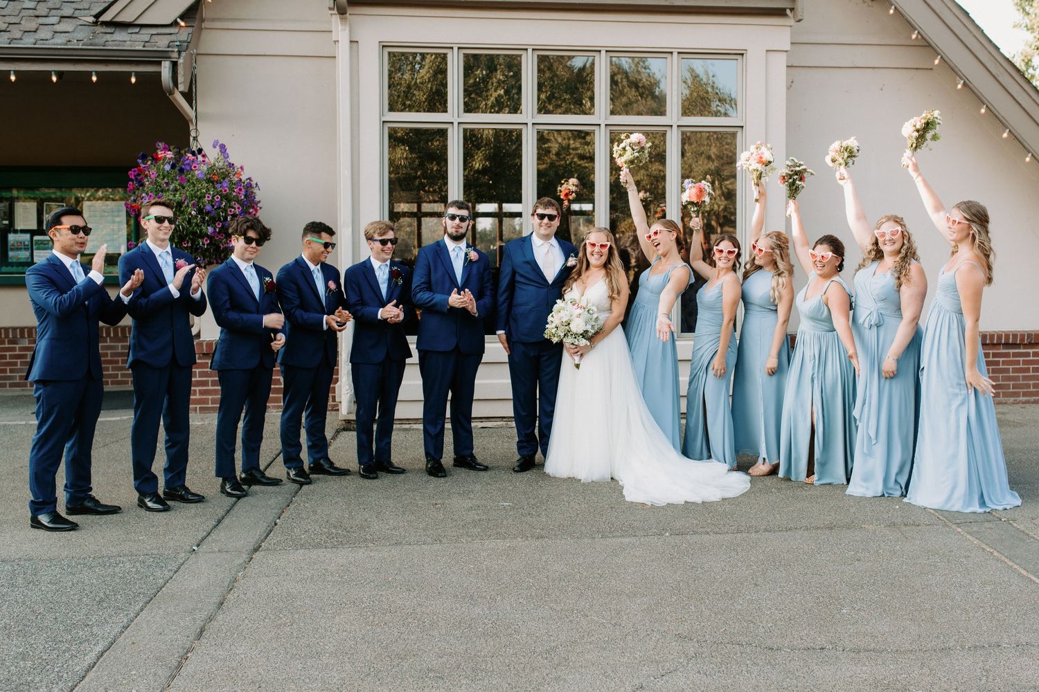 Wedding party in blue dresses and navy suits poses in front of white building.