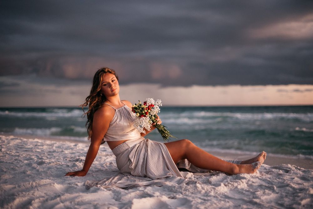Senior Portrait photographer captures a beautiful senior holding a bouquet of flowers while sitting on the white sandy beaches of Santa Rosa