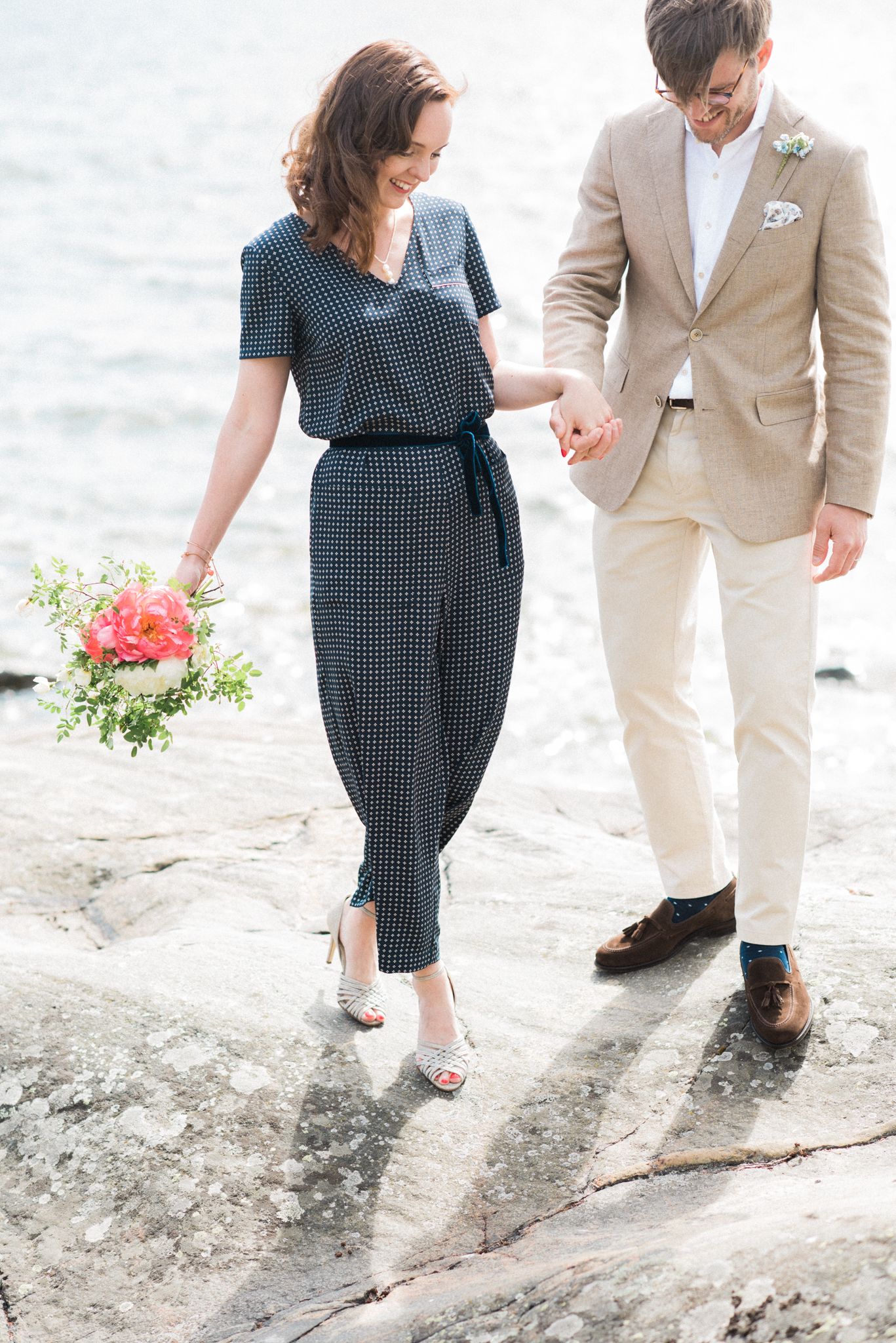 Couple walking on rocky shore with she wearing polka dot dress and he in beige suit holding a coral flower bouquet.