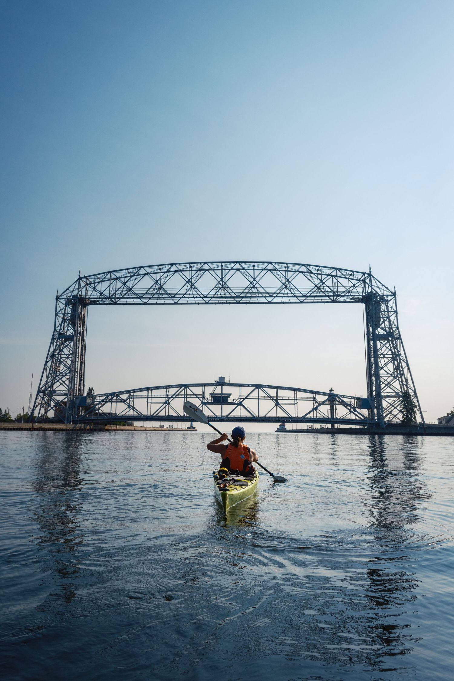 Kayaker paddling towards a large steel lift bridge over calm waters.