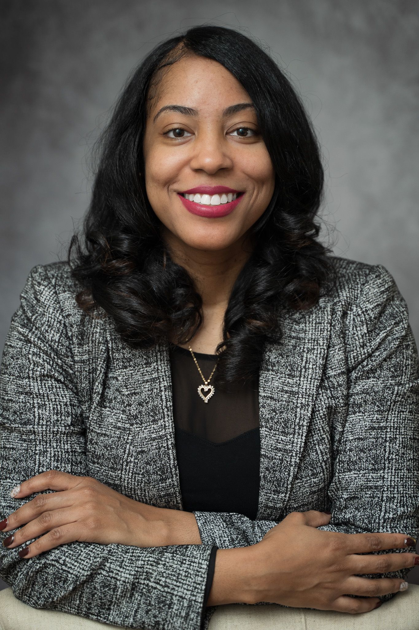 Professional headshot of person wearing grey blazer and black top with heart necklace.