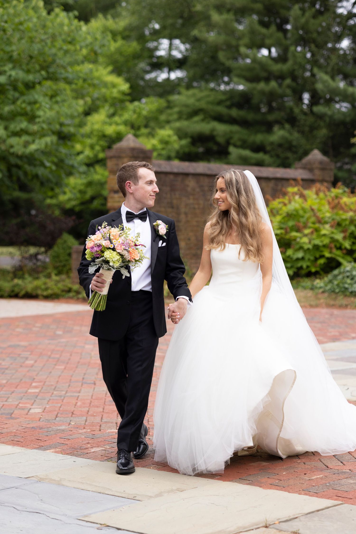 A bride in a white gown and groom in tuxedo walk hand-in-hand down a brick pathway surrounded by greenery.