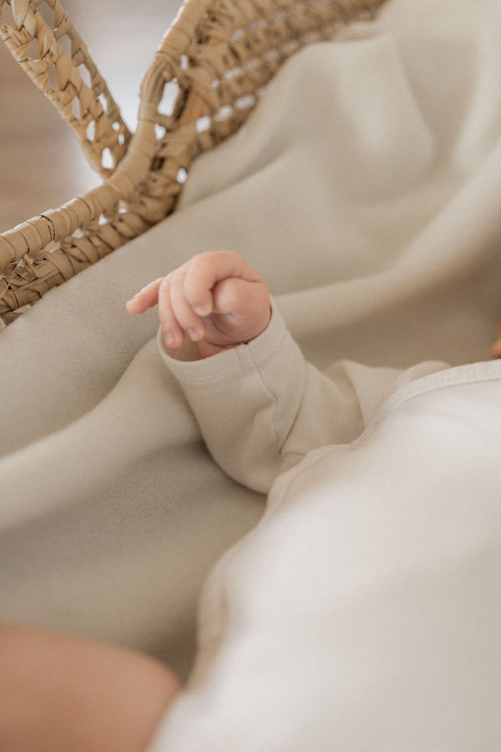 A small baby's hand rests on soft beige fabric next to woven basket details in a gentle, minimalist composition.