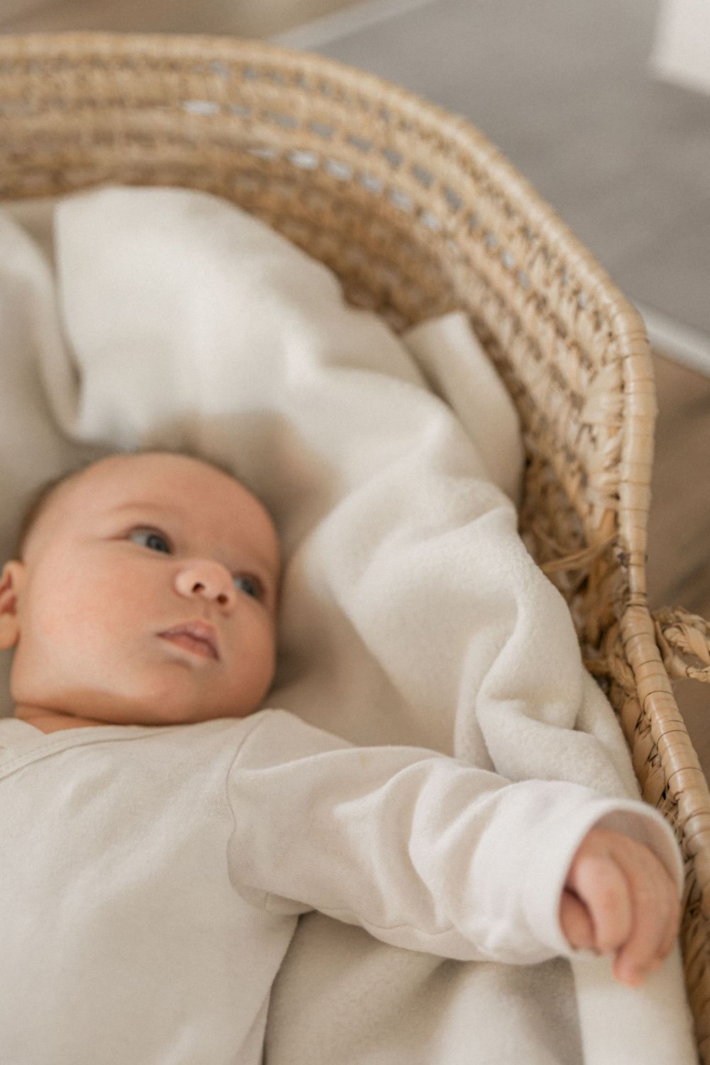 A baby in a cozy cream-colored outfit rests inside a woven wicker bassinet with soft white bedding.