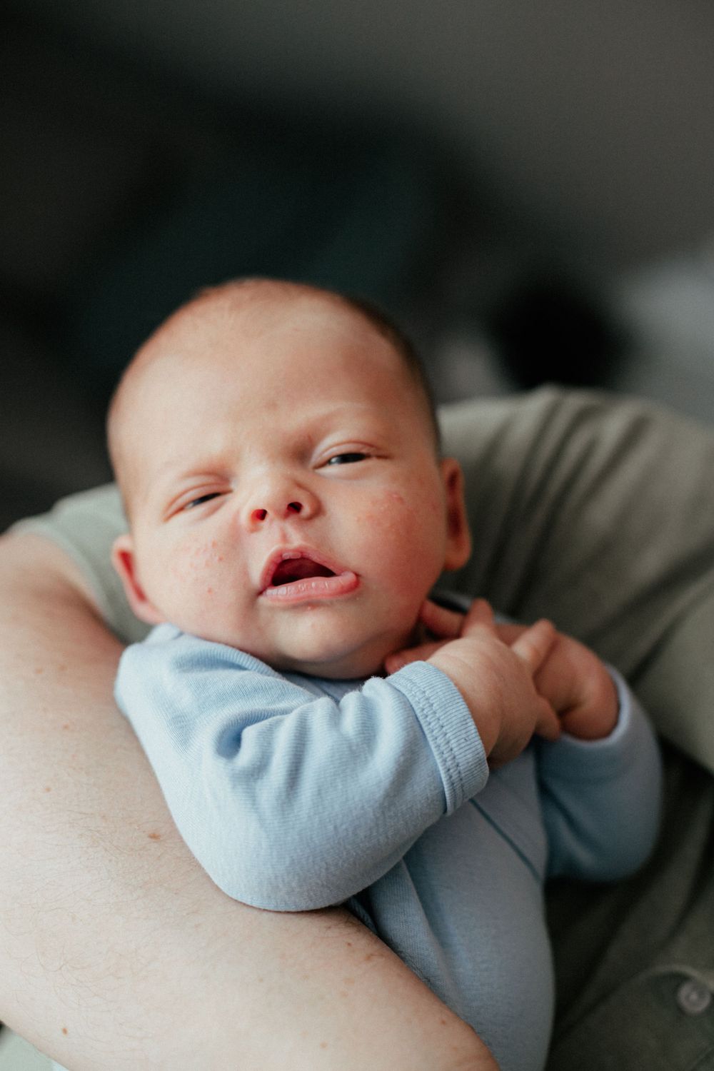 A sleeping newborn wearing a soft blue onesie rests peacefully against someone's arm in a dimly lit setting.