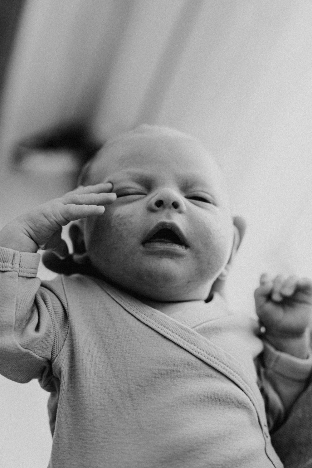 A close-up black and white photo of a yawning baby in a light onesie with tiny hands stretching upward.