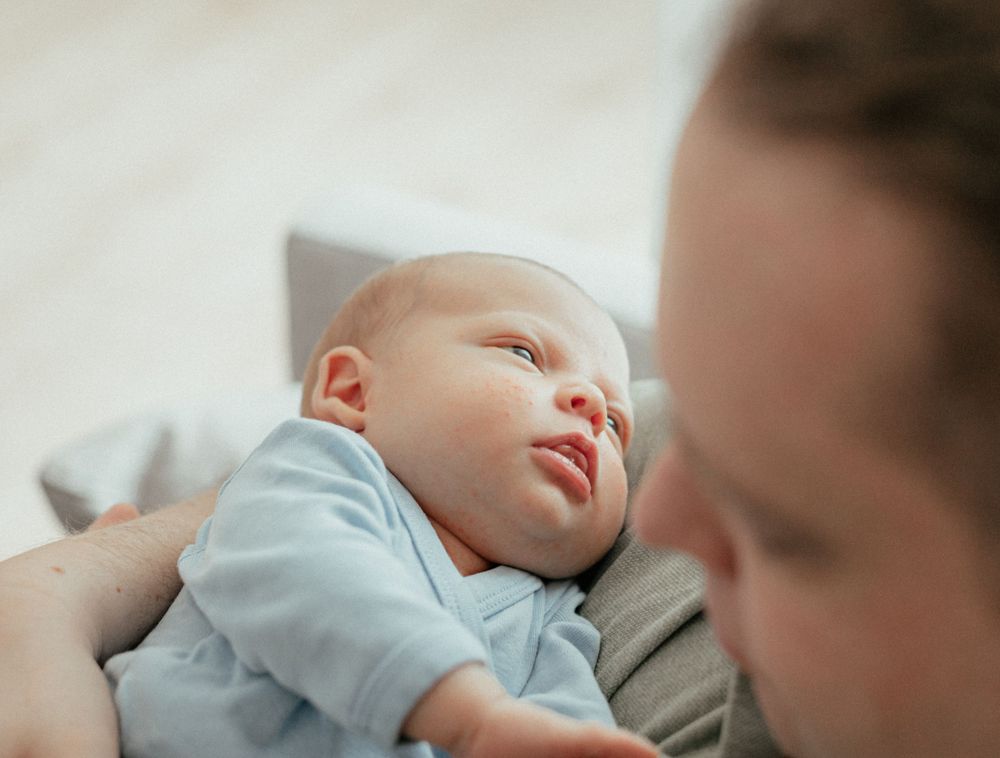 A peaceful baby in light blue clothing lies on soft bedding looking upward with gentle lighting in the background.