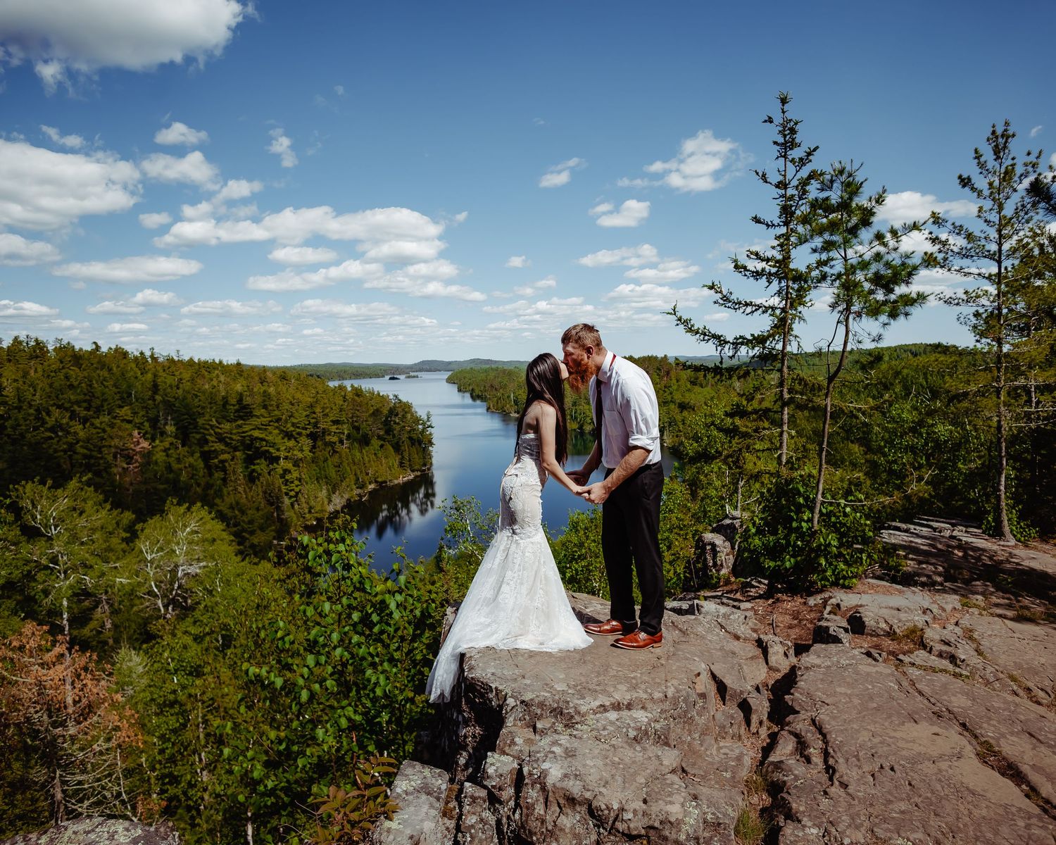 A wedding couple poses on a rocky cliff overlooking a vast forest landscape and winding river. Elopements Minnesota