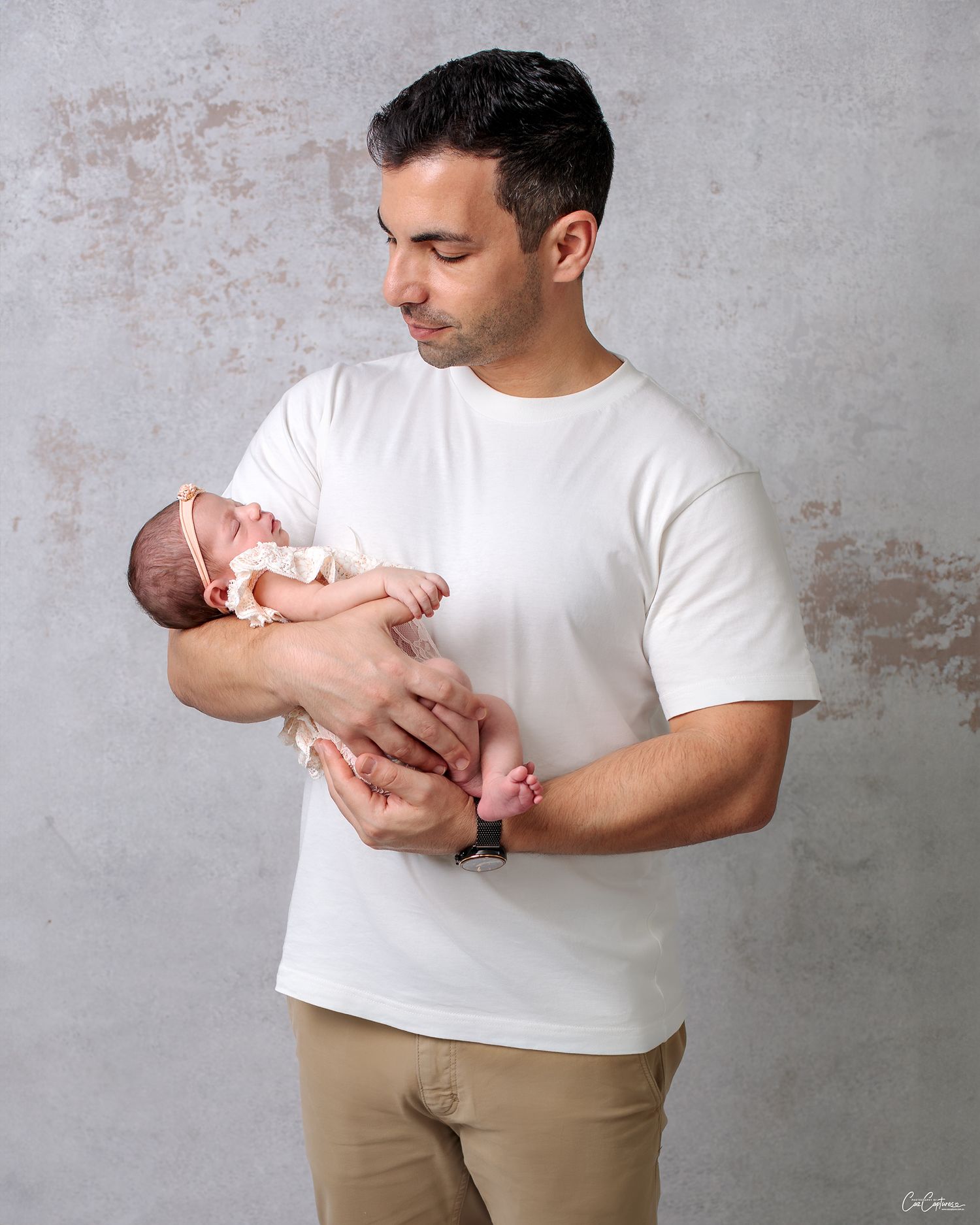 Fatherhood Newborn Photography of a dad lovingly holding his newborn daughter smiling proudly, wearing white against grey canvas backdrop.