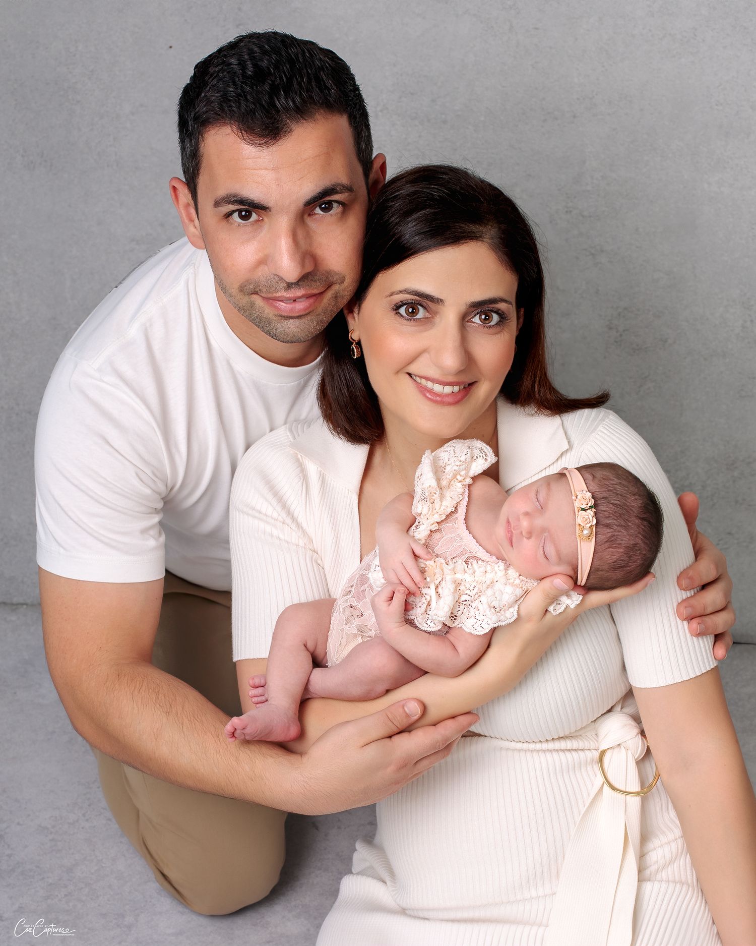 Mother and father newborn portrait, holding their baby girl proudly and lovingly smiling at the camera while the baby sleeps in their arms.