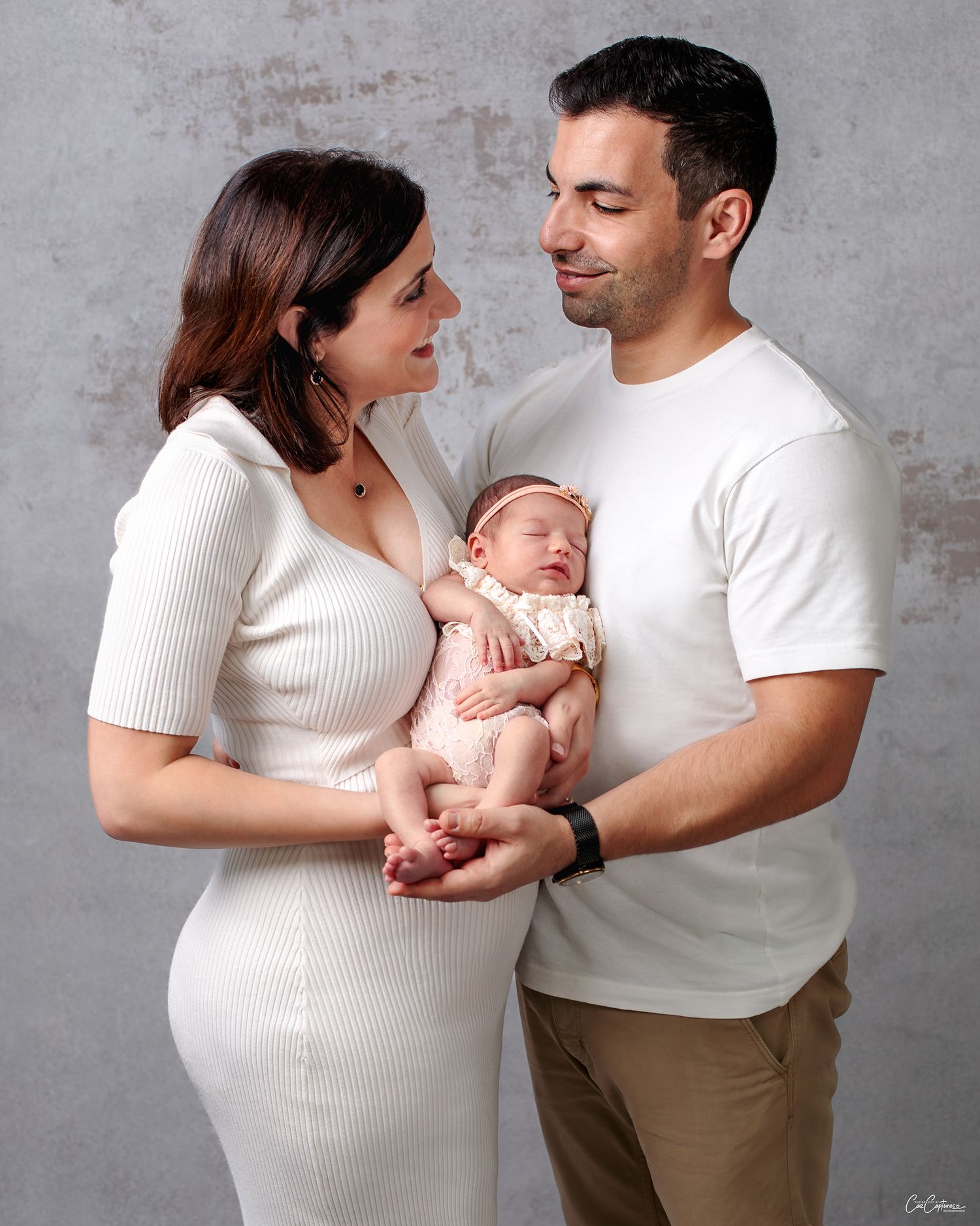 Newborn Family Portrait, Mother and Father holding their newborn baby girl between them looking lovingly at each other by Caz Captures.