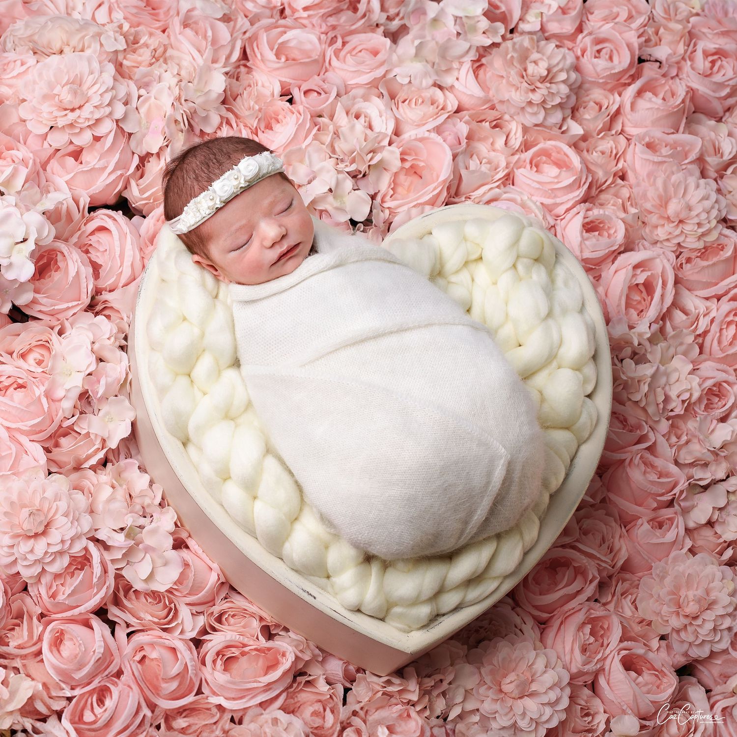 Stunning newborn photo of baby girl wrapped in white blanket on a white wooden heart against peach roses. The baby girl is sleeping.
