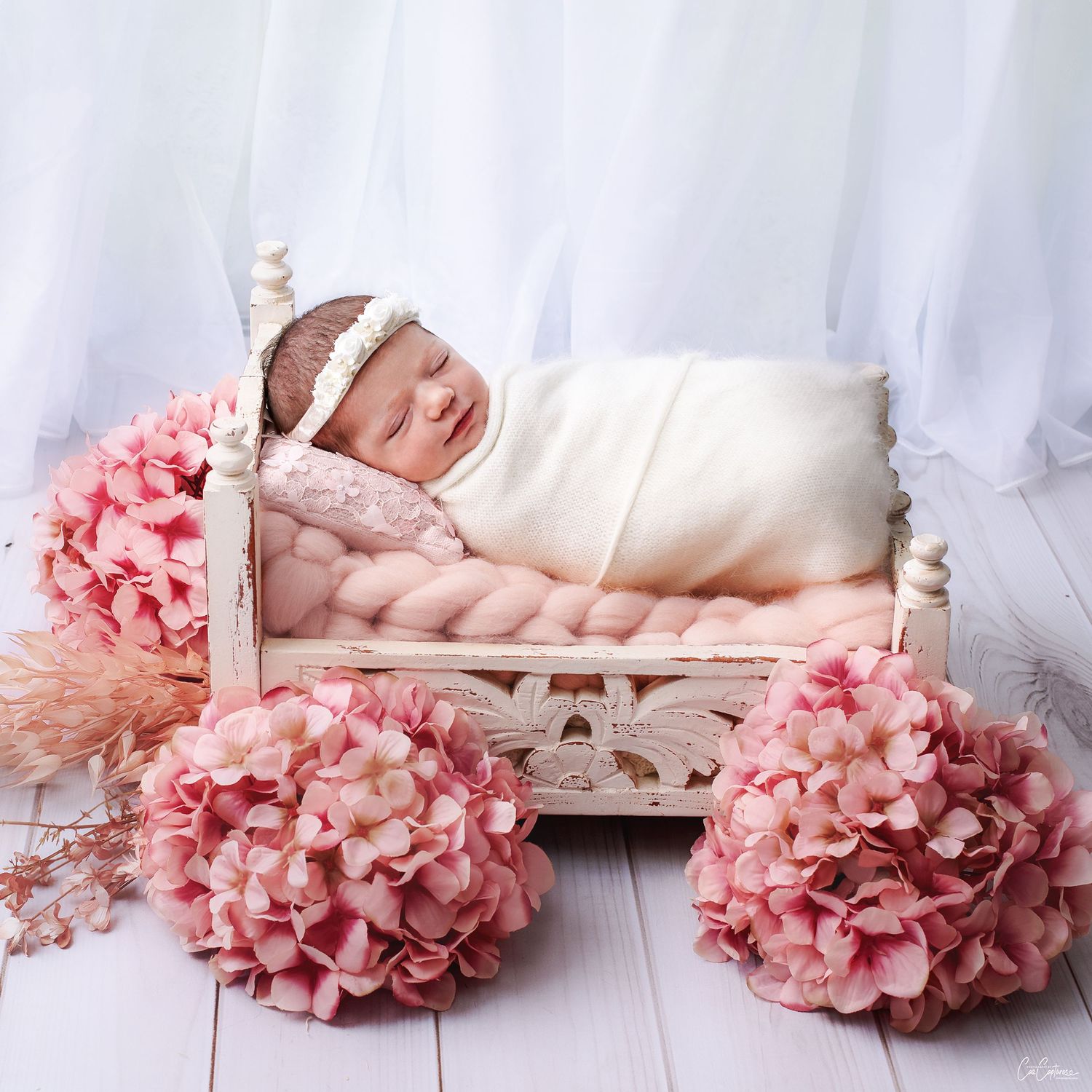 Beautiful newborn photography of baby girl sleeping on a tiny white ornate bed surrounded by pink hydrangeas against sheer white curtains.