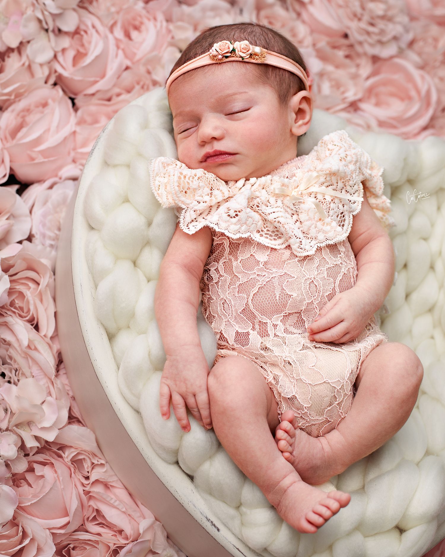 Close up of newborn baby girl sleeping peacefully wearing beautiful peach coloured lace romper and matching headband on white wooden bowl.