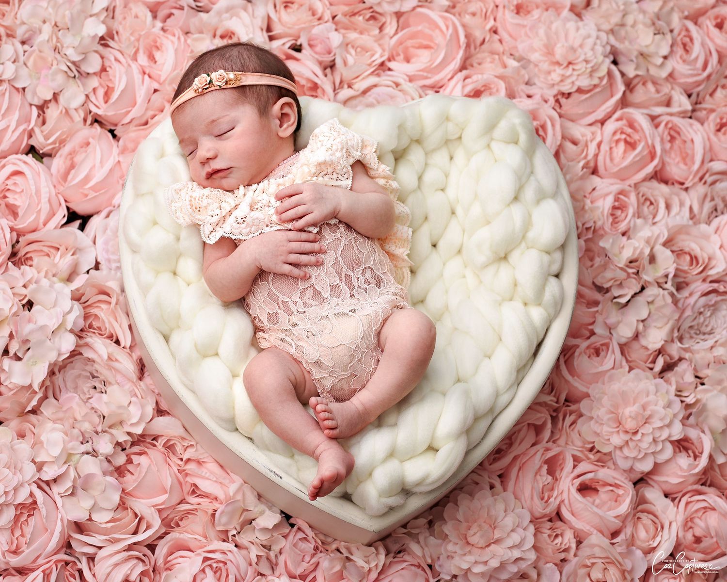Stunning newborn photo of baby girl in peach lace romper on a white wooden heart against peach roses. The baby girl is sleeping.
