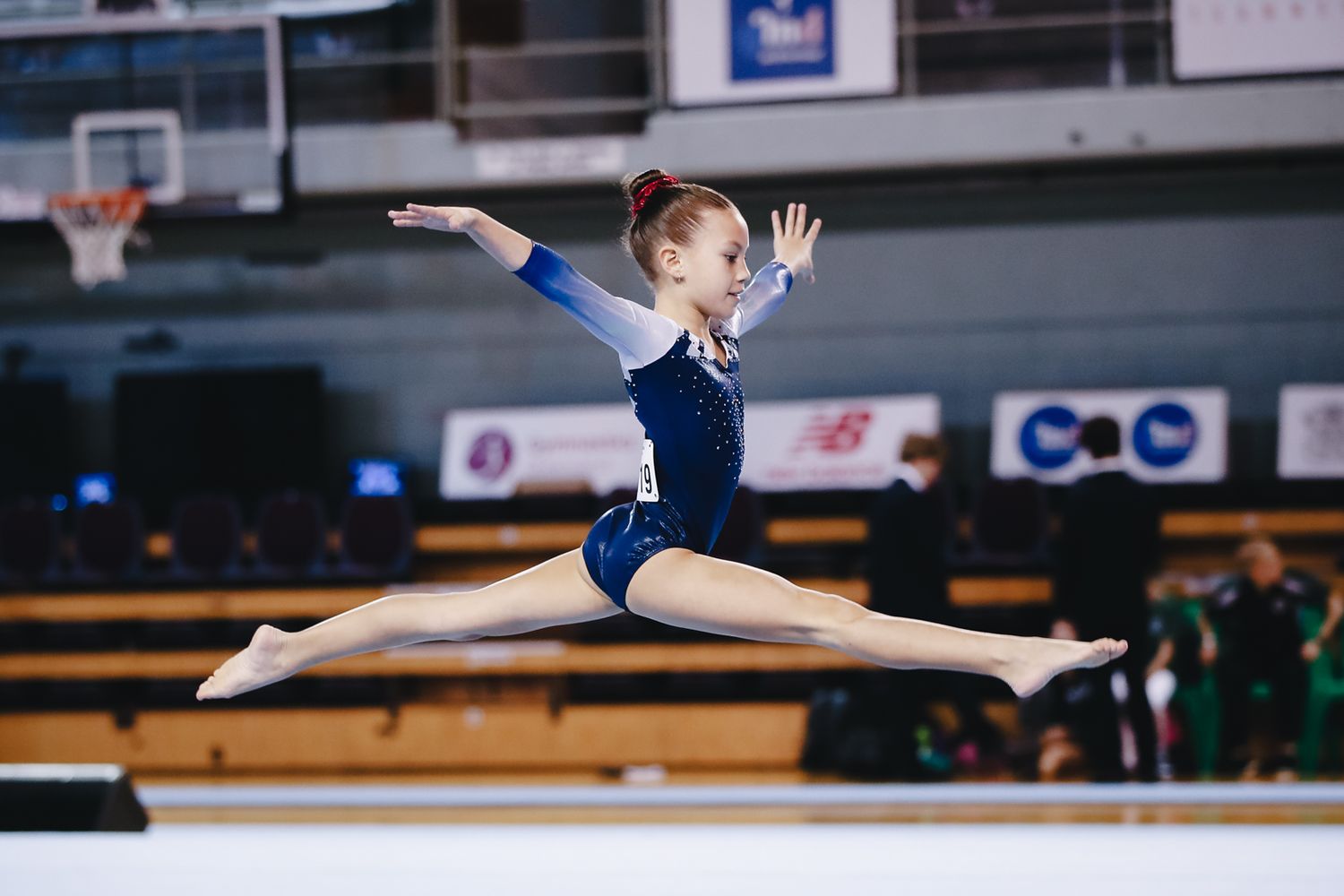 Gymnast in blue leotard executes perfect split leap during floor routine.