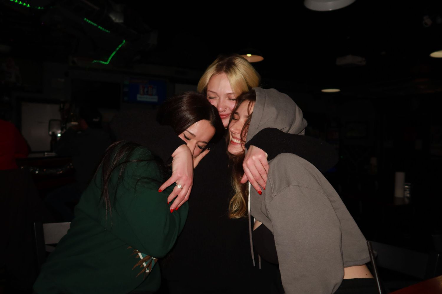 Three young women hugging and smiling together in a dimly lit venue at night.