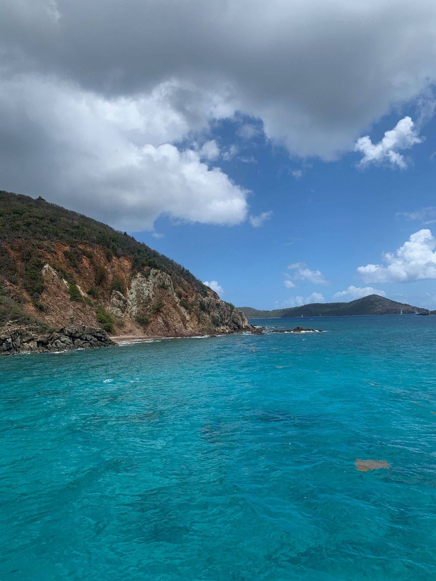 Turquoise Caribbean waters surround a rocky hillside under a partly cloudy blue sky in a tropical island paradise.