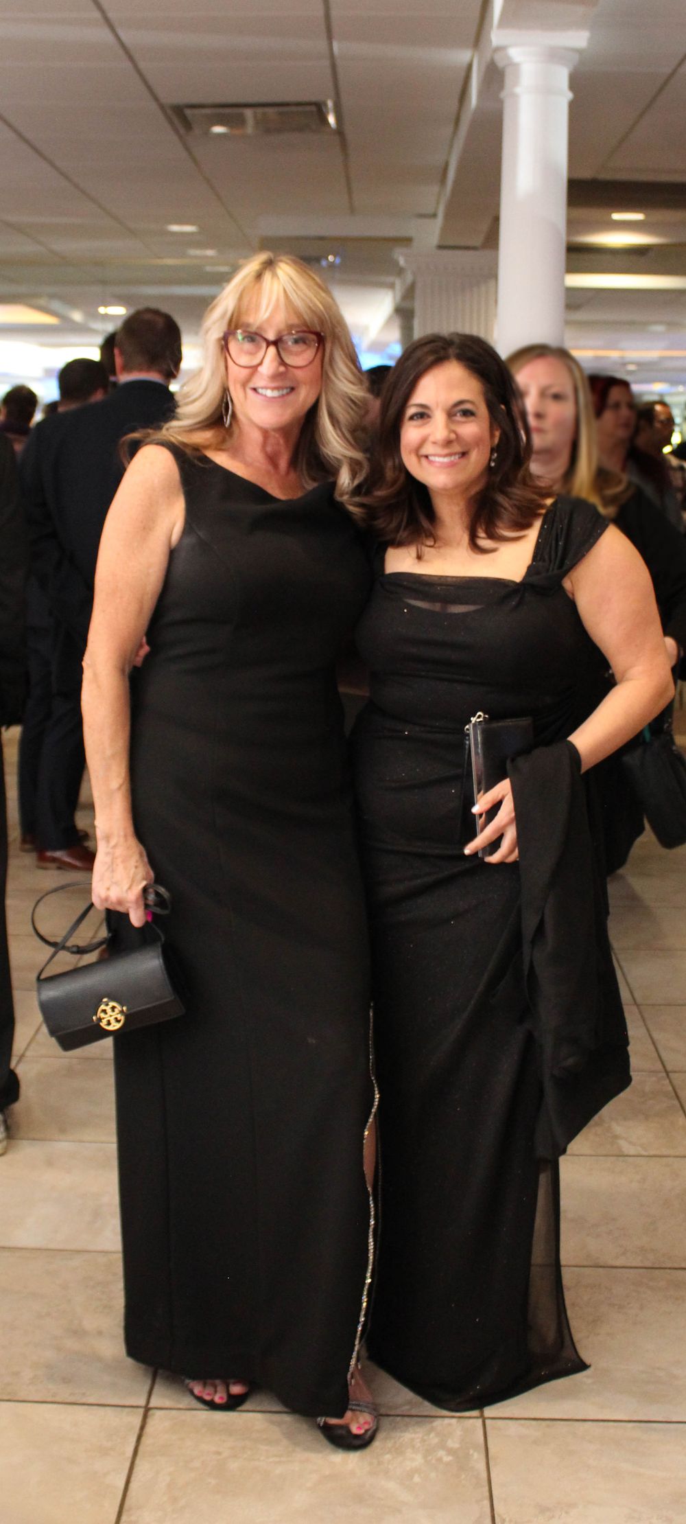 Two women wearing elegant black evening gowns pose together at a formal event.