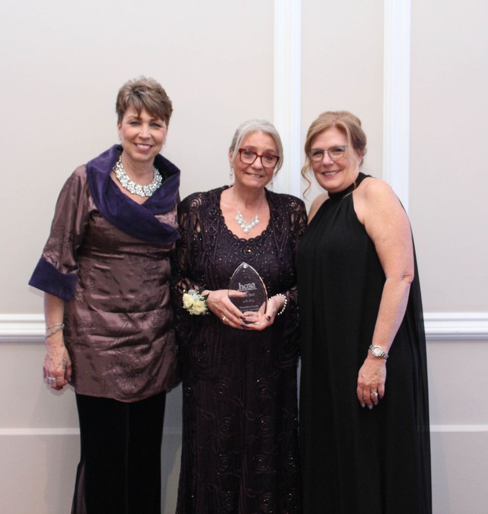 Three people in formal attire pose together at an elegant evening event with white wall panels.