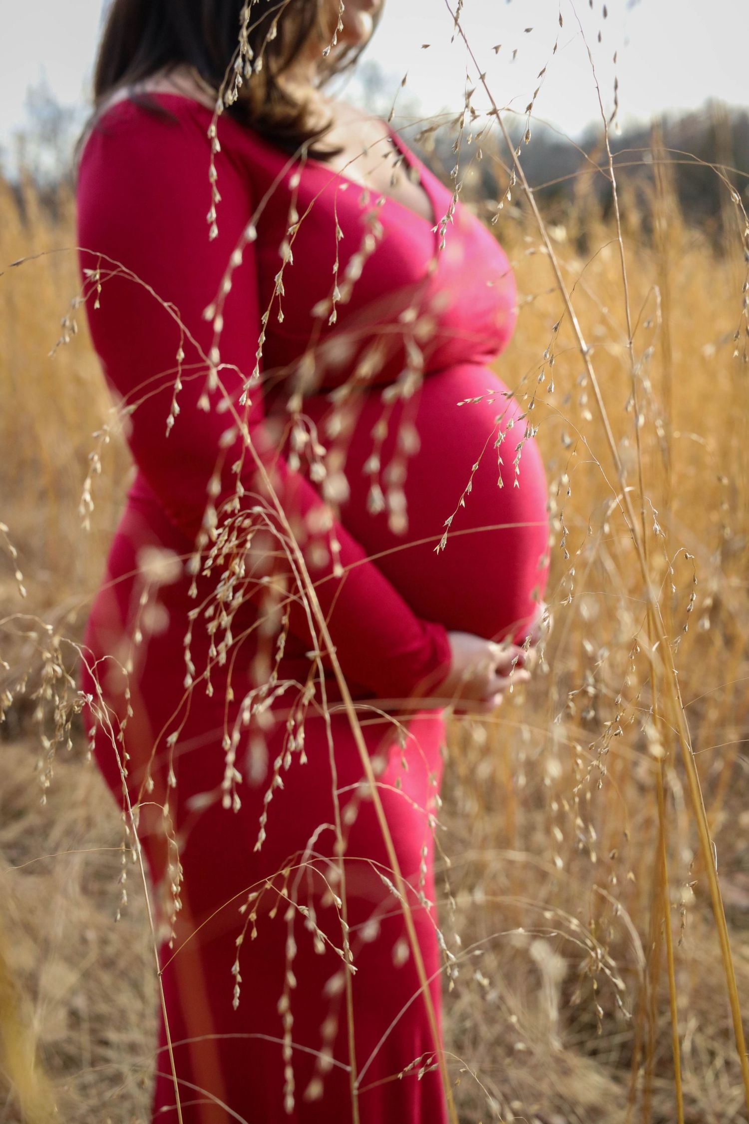 Woman in red dress standing in golden wheat field during maternity photoshoot.