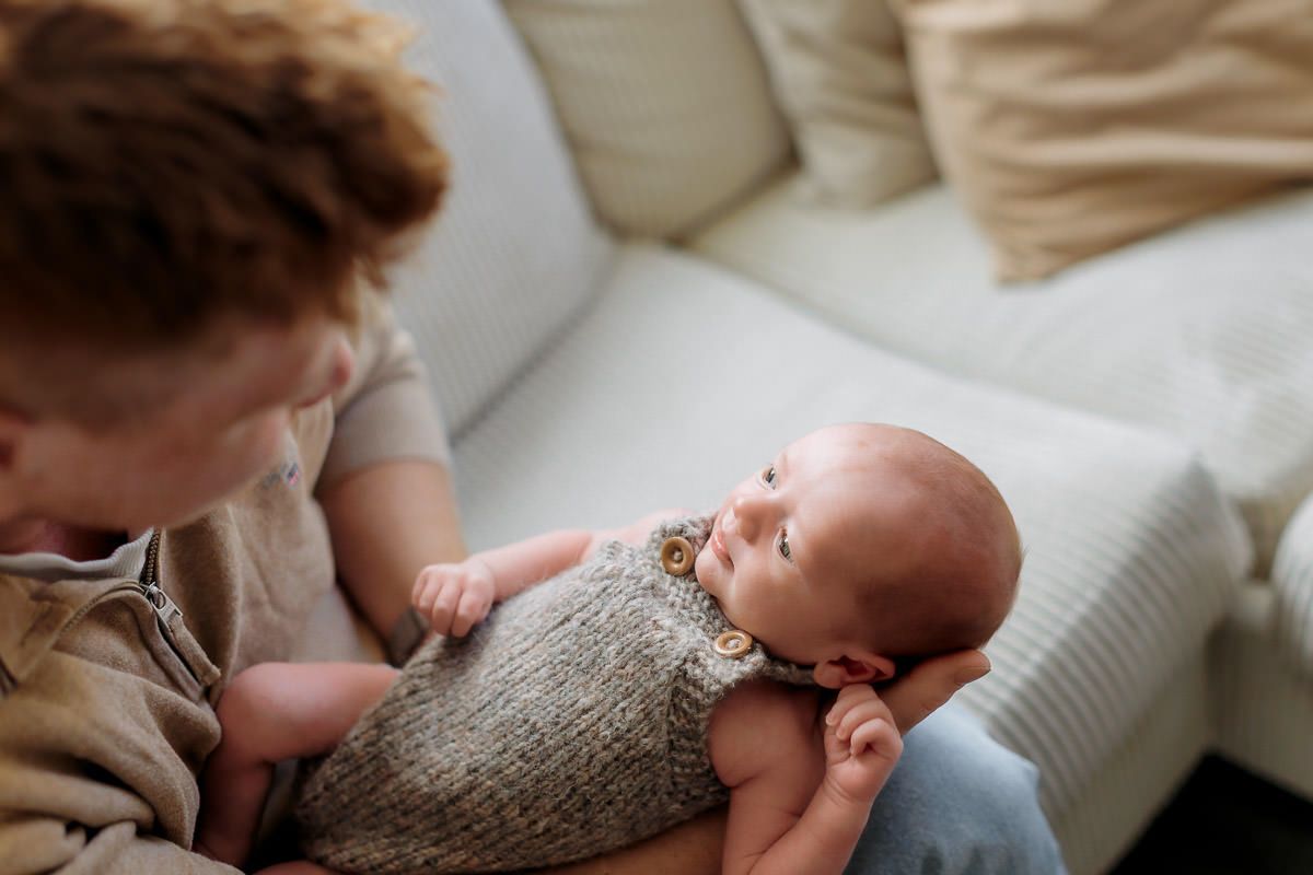 Nyfødt gutt ligger i armene til pappa i sofaen og ser opp på han.