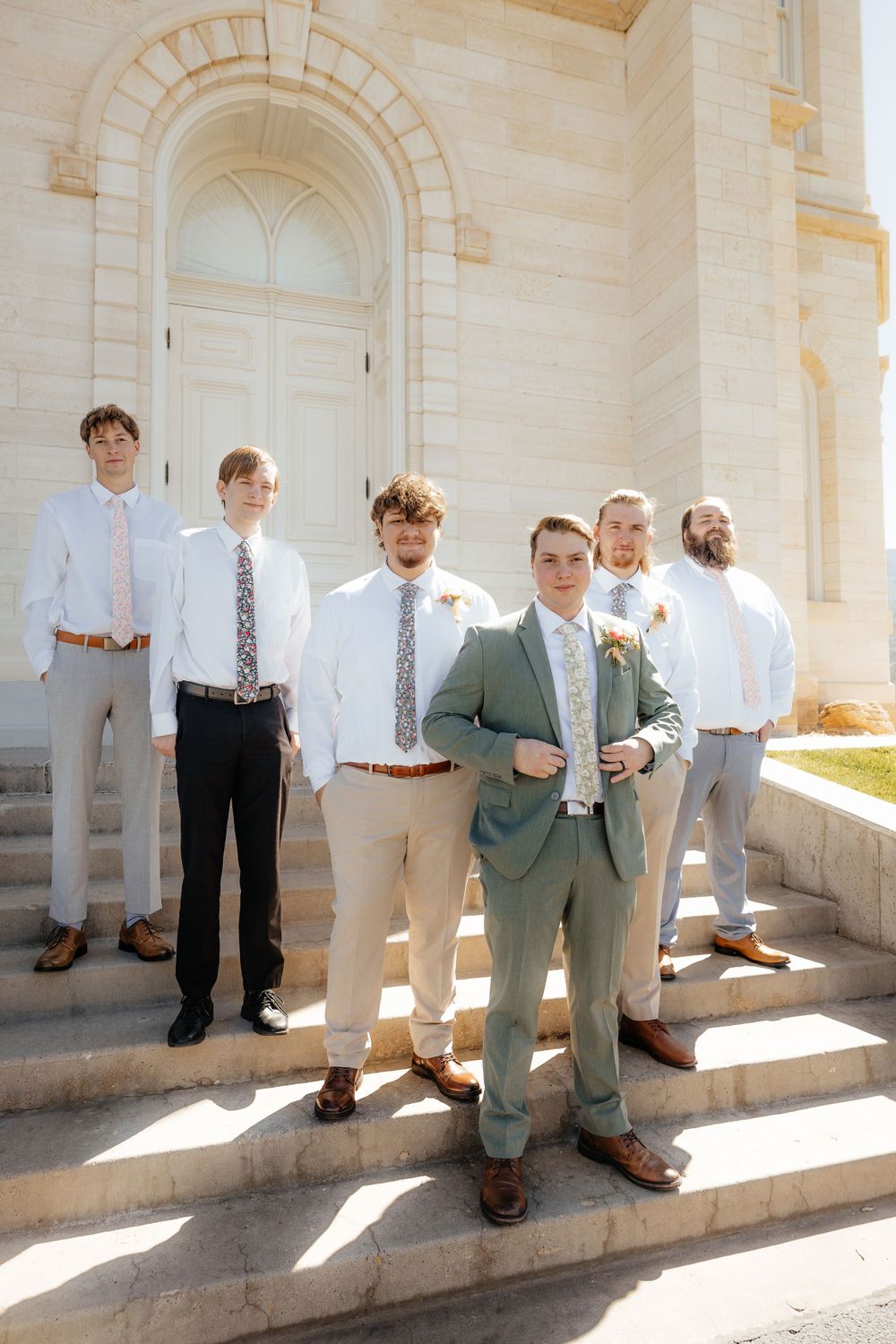 Groom in sage green suit stands with groomsmen in white shirts on church steps in sunny outdoor wedding photo.