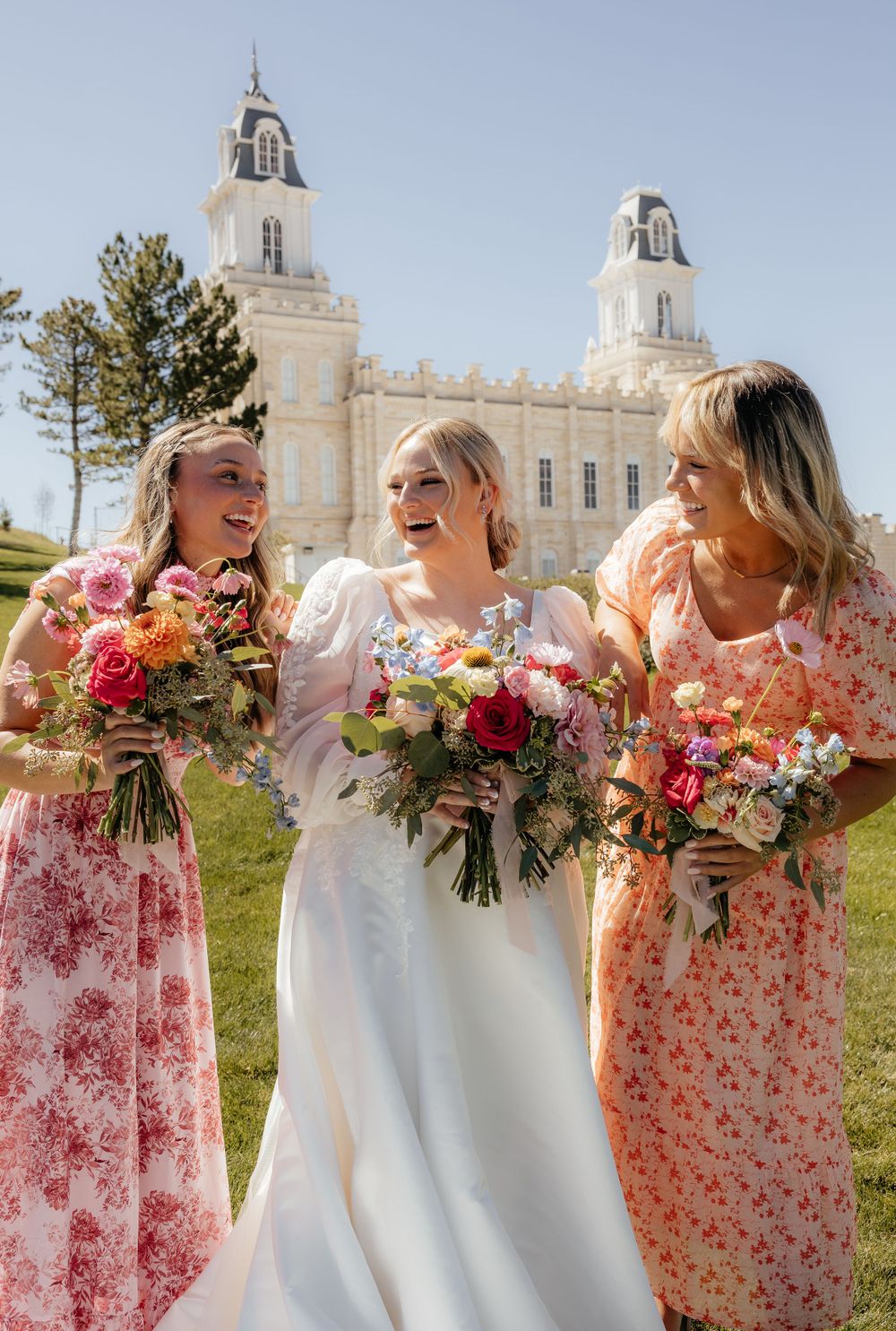 Bride and two bridesmaids in floral dresses laugh together holding colorful bouquets before a temple.