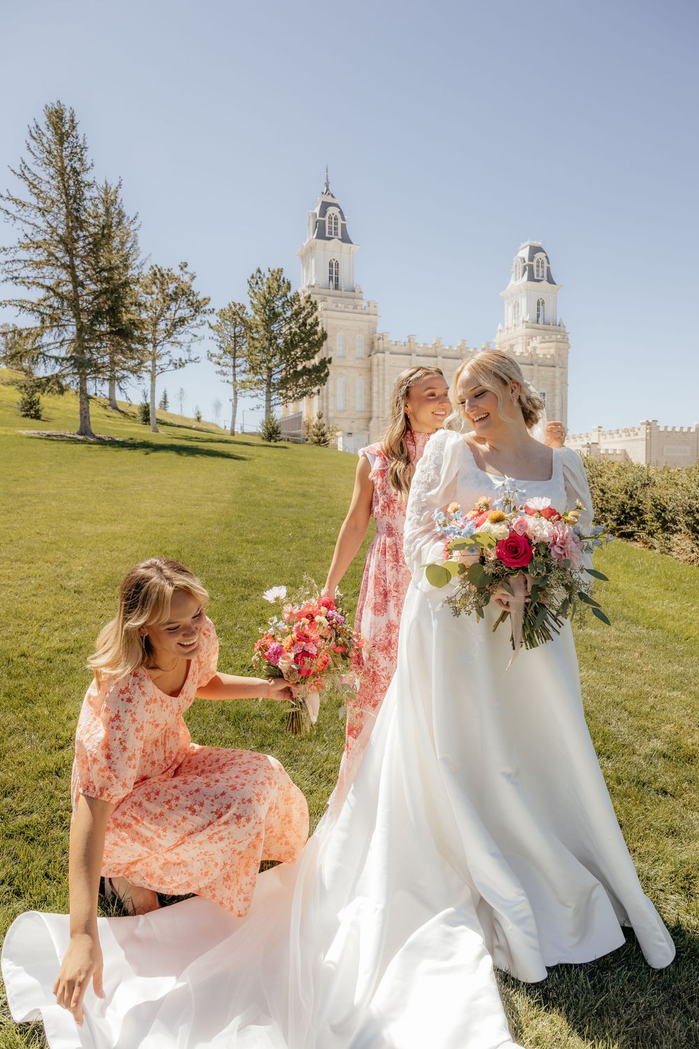 Bride in white gown holds colorful bouquet with two bridesmaids in pink floral dresses outside a temple.