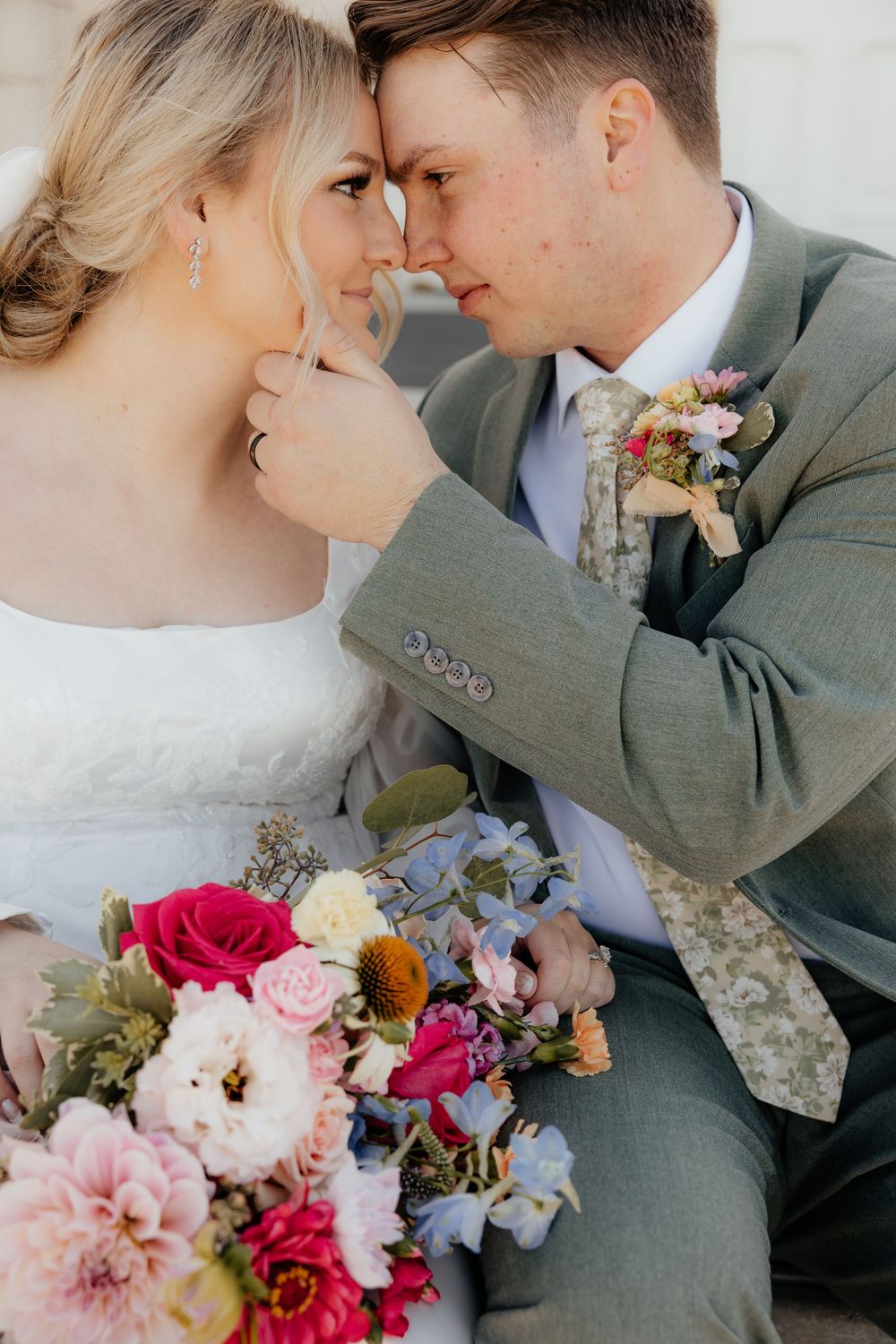 Bride and groom share an intimate forehead touch moment, holding a colorful bridal bouquet of pink and blue flowers.