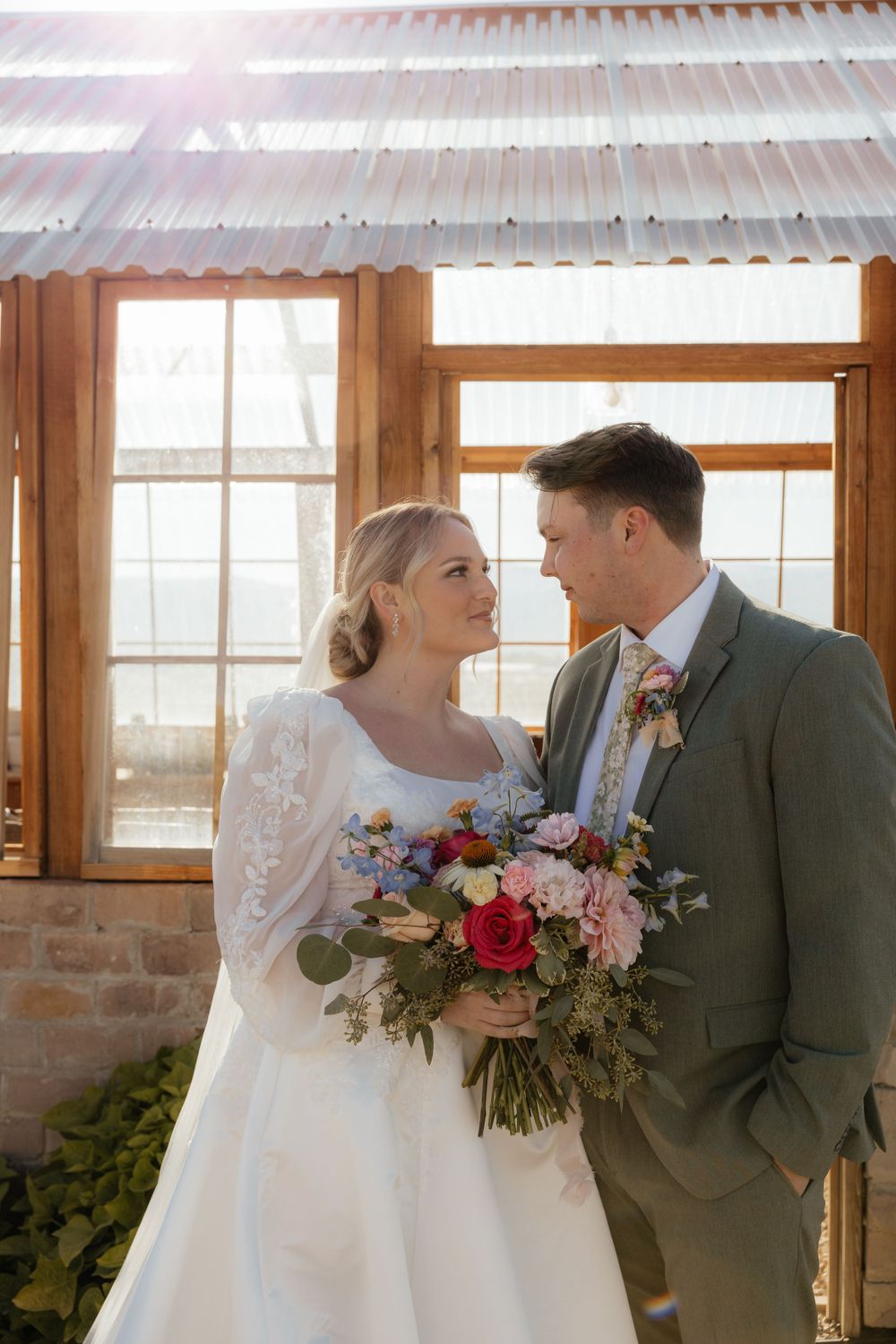 Bride and groom share a loving gaze holding a colorful bouquet inside a rustic sunlit greenhouse venue.