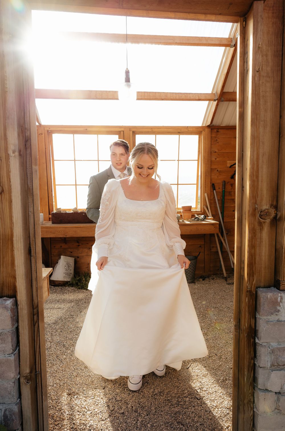 Bride in white lace gown smiles while lifting her dress hem inside a rustic sunlit wooden venue.