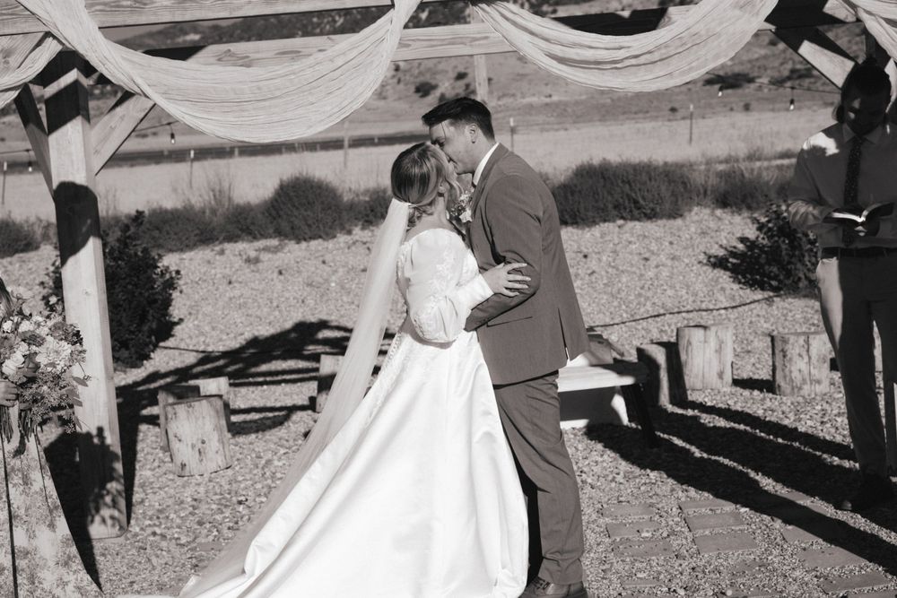 Bride and groom sharing their first kiss at an outdoor wedding ceremony beneath a rustic wooden arch.