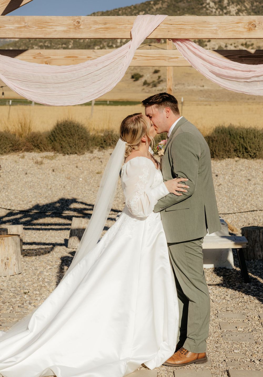 Bride and groom share first kiss under rustic wooden arch with pink draping at outdoor ceremony.