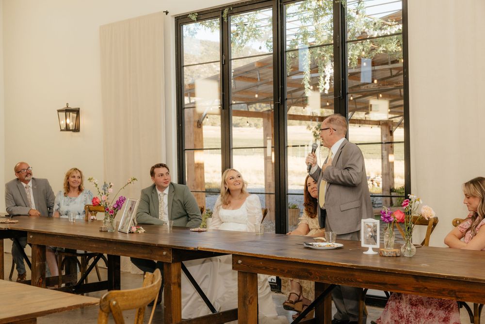 Man giving toast at rustic wedding reception with bridal party seated at long wooden farm table.